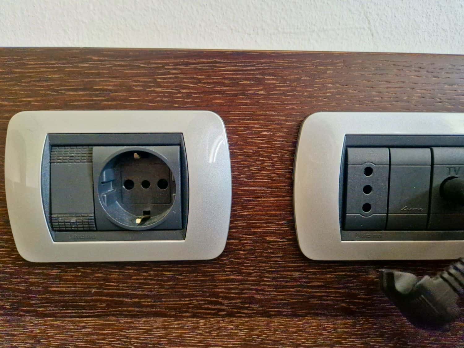 A close-up of two wall sockets on a wooden surface. The left socket is a European-style power outlet, and the right socket includes connectors for TV and radio. A black power plug is partially inserted into the TV socket.