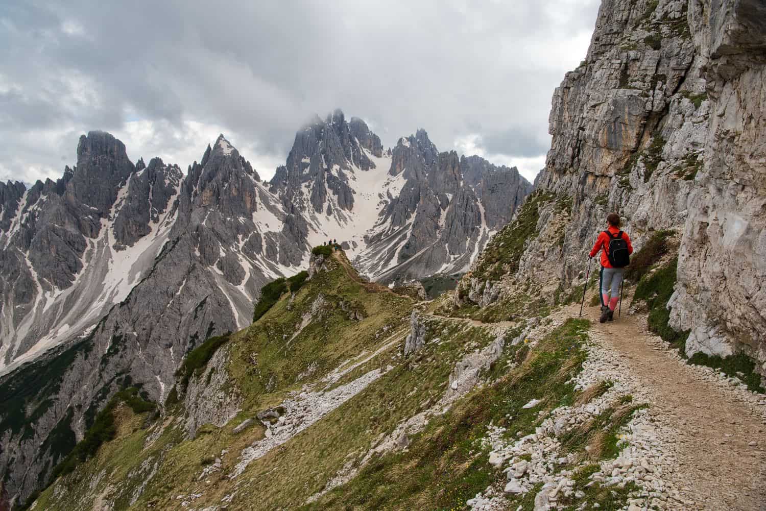 A hiker in a red jacket walks along a narrow mountain trail, surrounded by rugged rocky peaks partially covered in snow. The sky is cloudy, creating a dramatic backdrop for the towering mountains.