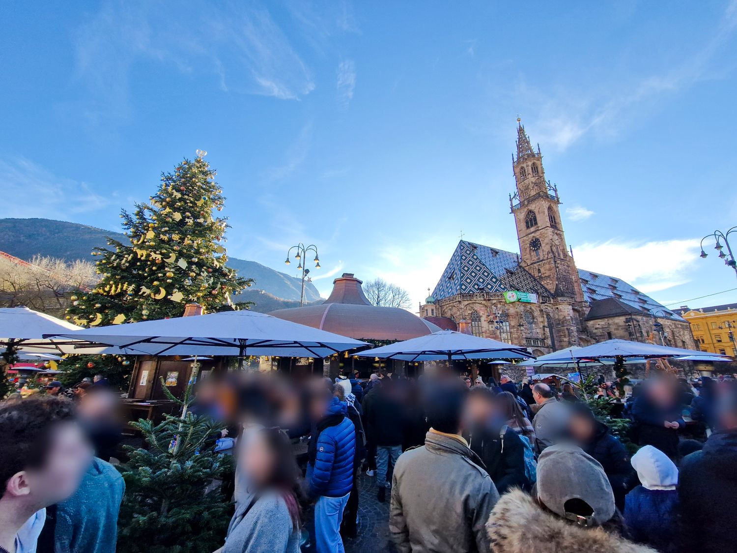 A crowded Christmas market fills a town square with people standing beneath white vendor umbrellas and evergreen decorations. A large decorated Christmas tree stands to the left, while a historic church with a patterned roof and tall bell tower rises behind the stalls under a clear blue sky. The image shows a festive winter atmosphere in a European city center.