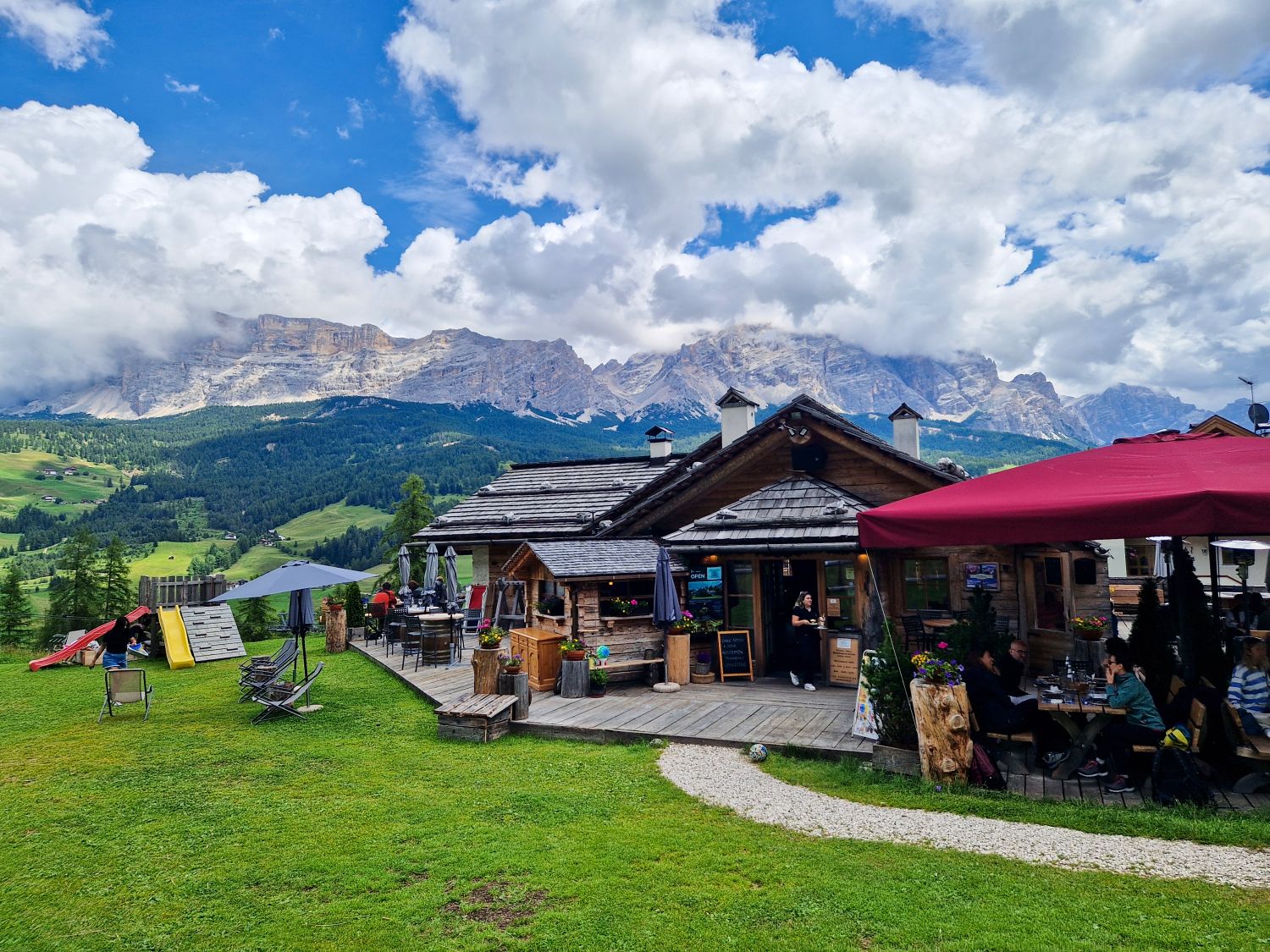 A rustic wooden mountain restaurant sits on a grassy hillside with outdoor tables and people dining under a red canopy. Rolling green fields lead to dramatic rocky mountains in the background, partially covered by large white clouds. The image shows a relaxed alpine setting combining food, nature, and scenic views.