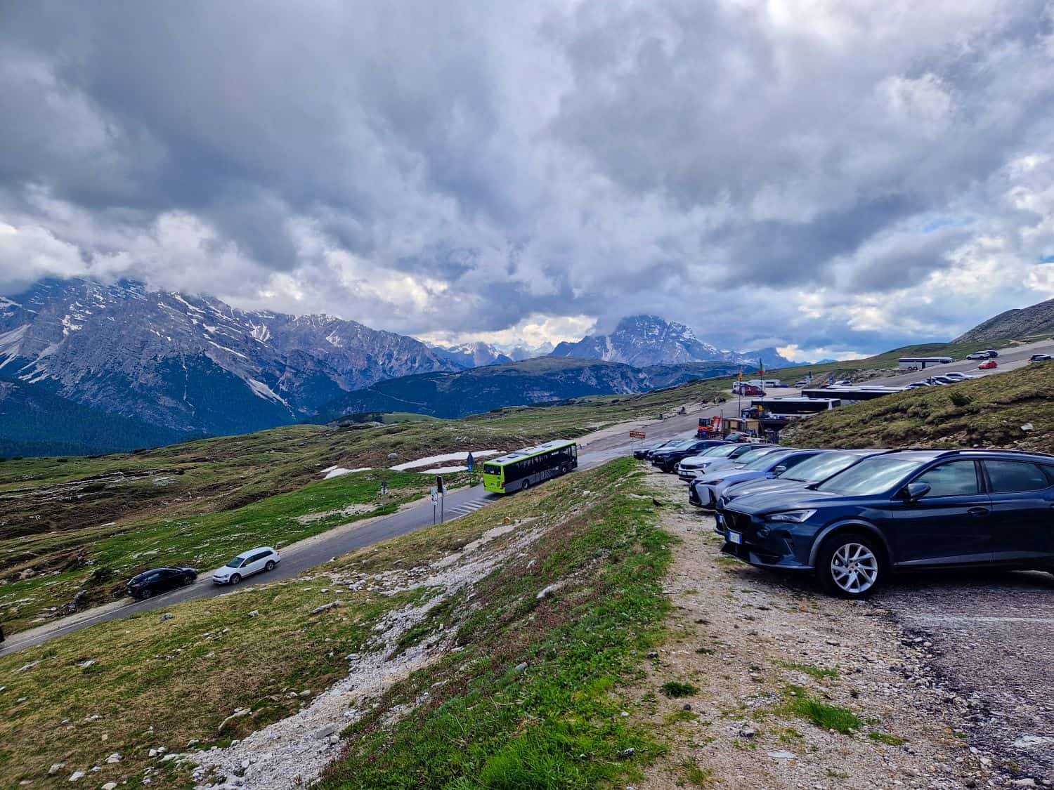 A parking area next to a mountain road with several cars parked. A green bus drives along the road. Snow-capped mountains and a cloudy sky form the background, with patches of green grass on the slopes.