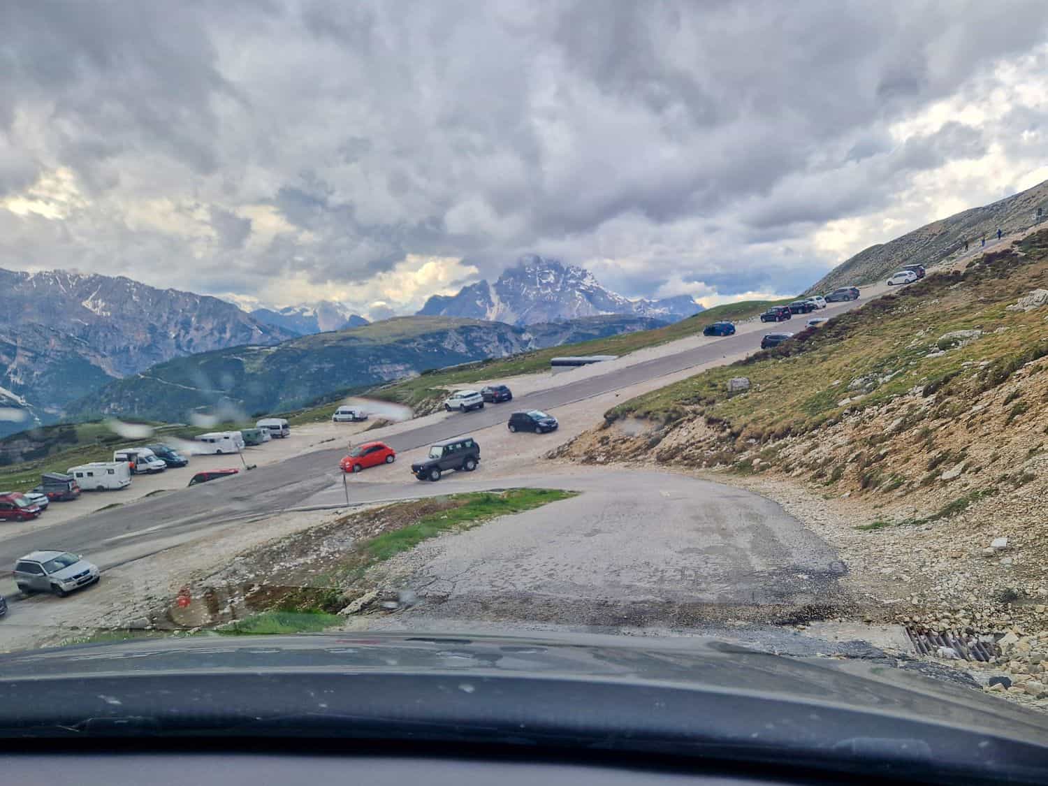 A scenic mountain road with multiple cars parked along its side, winding through rocky terrain. The sky is overcast, with dramatic clouds, and distant mountain peaks are visible in the background, partly covered in snow.