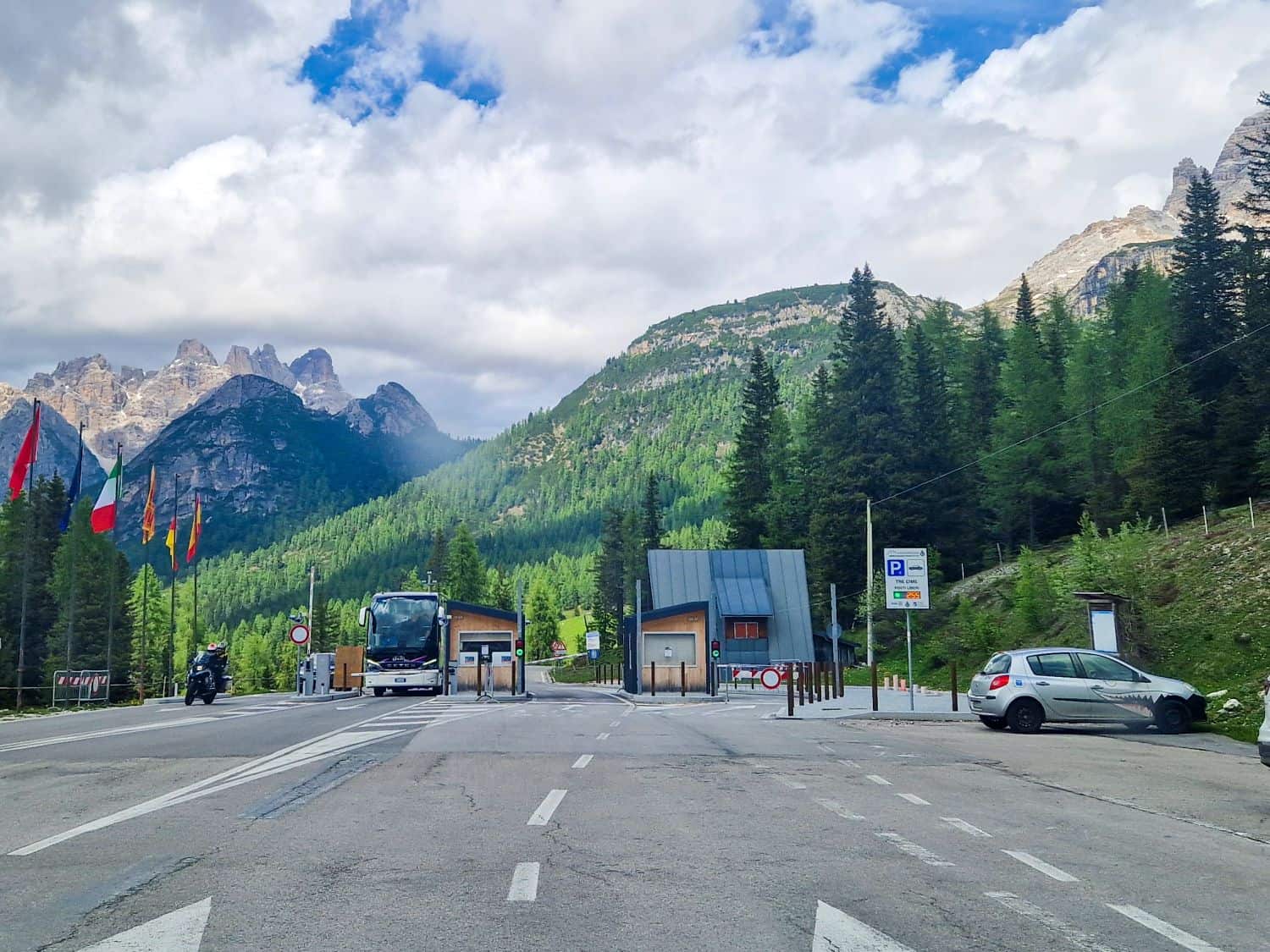 A scenic mountain road with a bus station. A bus is parked to the left, and a car is on the right. Flags line the road, with lush green trees and rugged mountains in the background under a partly cloudy sky.