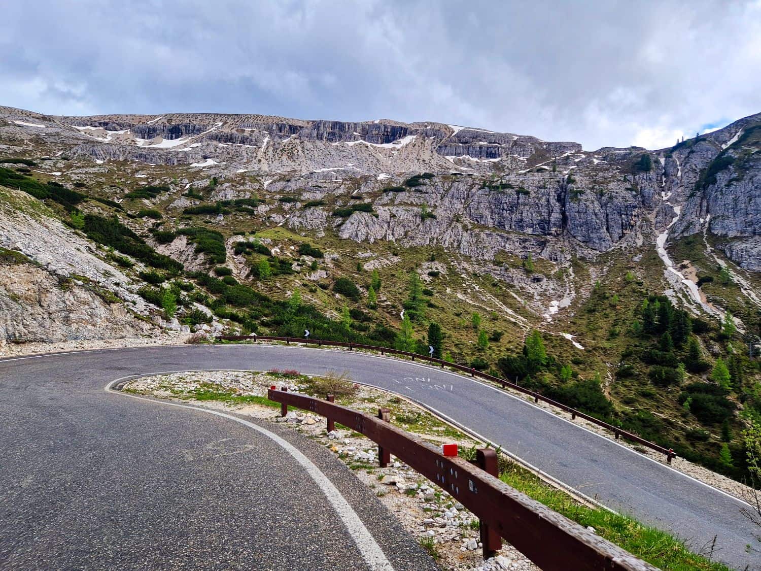 Mountain road with sharp curves winding through rocky terrain and sparse greenery under a cloudy sky. Rugged cliffs rise in the background. Red and white painted guardrails line the road, providing protection on the sharp turns.