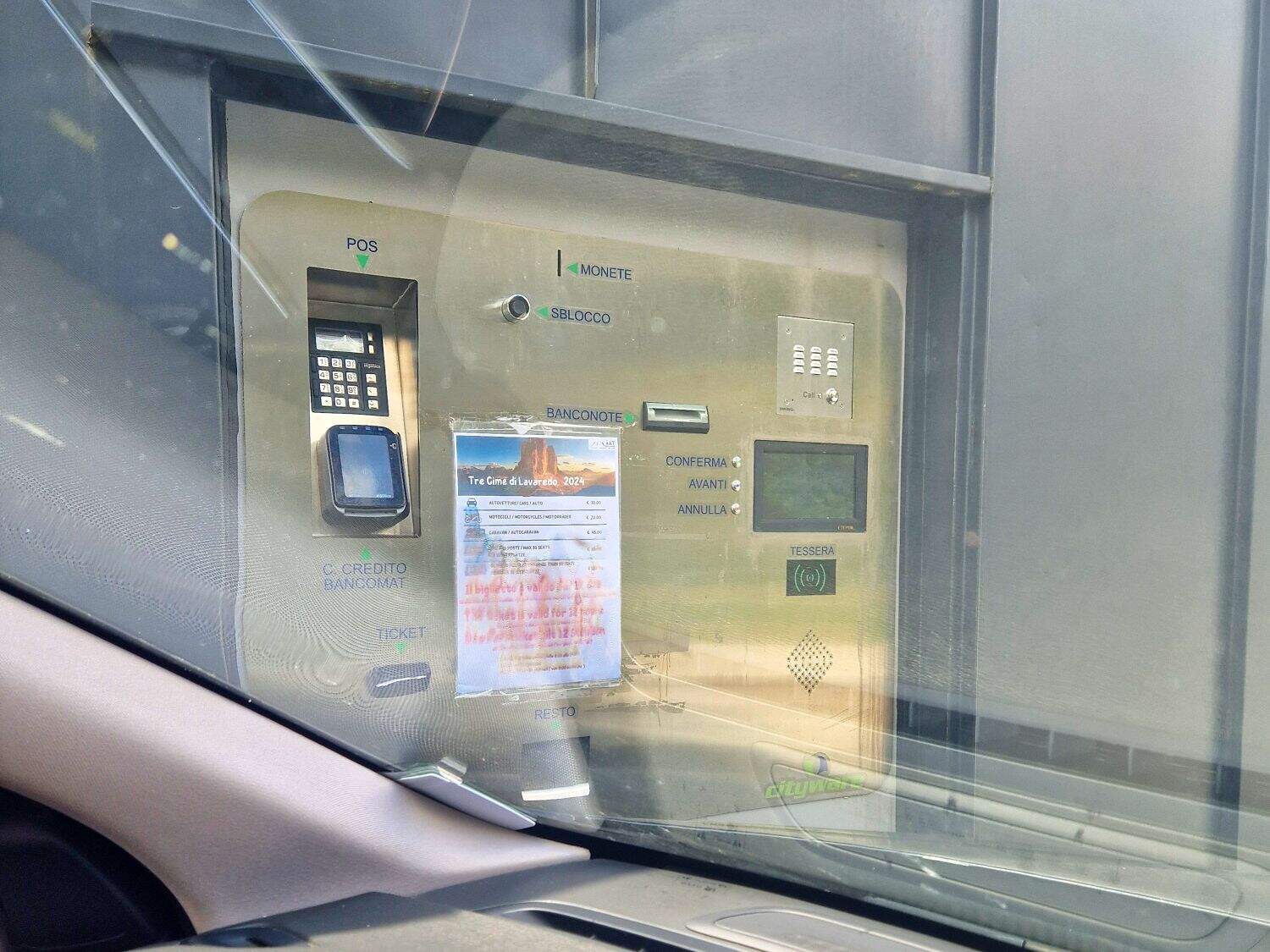 A parking payment machine viewed from a car window. It includes card and cash slots, a keypad, and a screen. A partially obscured poster is taped to it. The surroundings appear to be an indoor parking facility.
