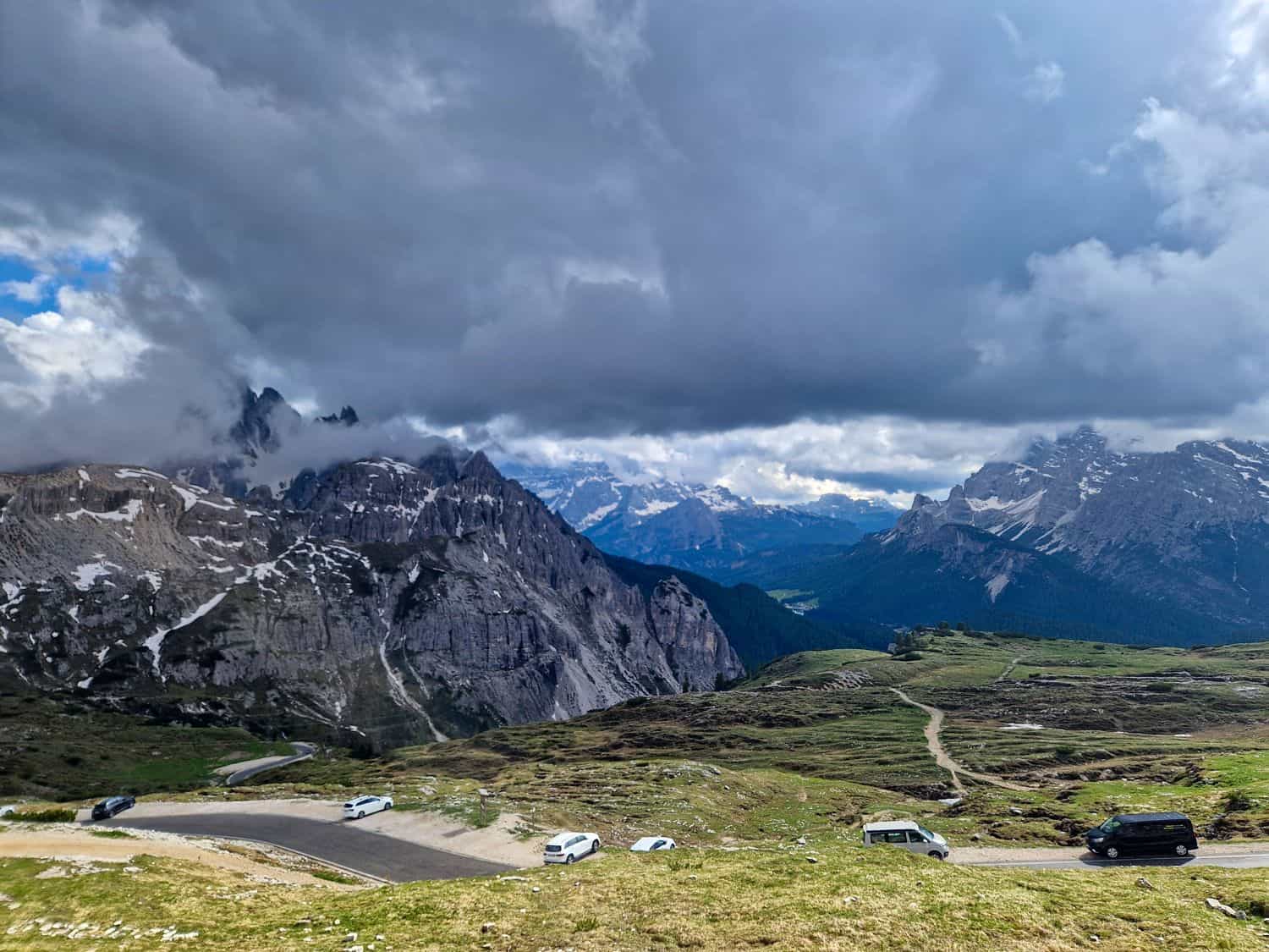 A scenic view of a mountainous landscape under dramatic clouds. Snow-capped peaks are partially covered by fog, with a winding road and several vehicles in the foreground. Green hills contrast with the rugged, rocky terrain.