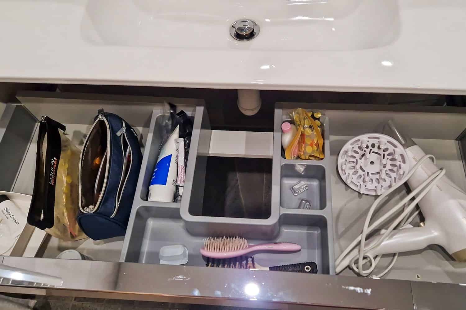 An open bathroom drawer beneath a sink, organized with various compartments. It contains toiletry bags, a tube of toothpaste, a hairbrush, a hairdryer, and small containers. The drawer is neatly arranged for easy access to personal care items.