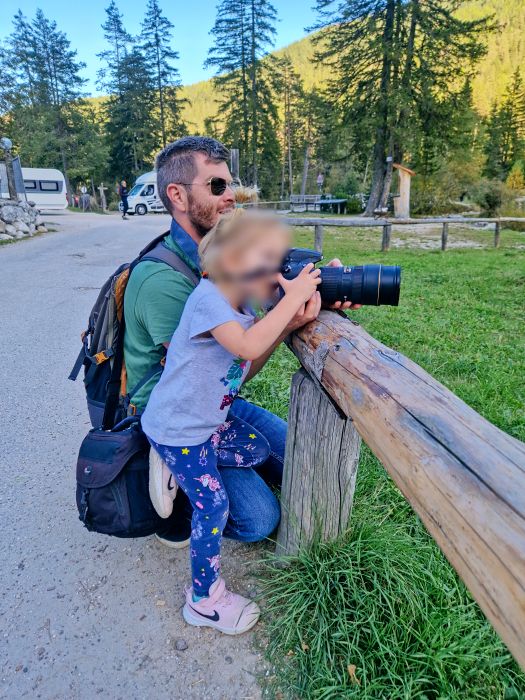 Lago di Braies: Discover The Pearl of the Dolomites 8 An adult kneels next to a child, helping them hold a large camera on a wooden fence in a park. Trees and a parked camper van are visible in the background under a sunny sky.