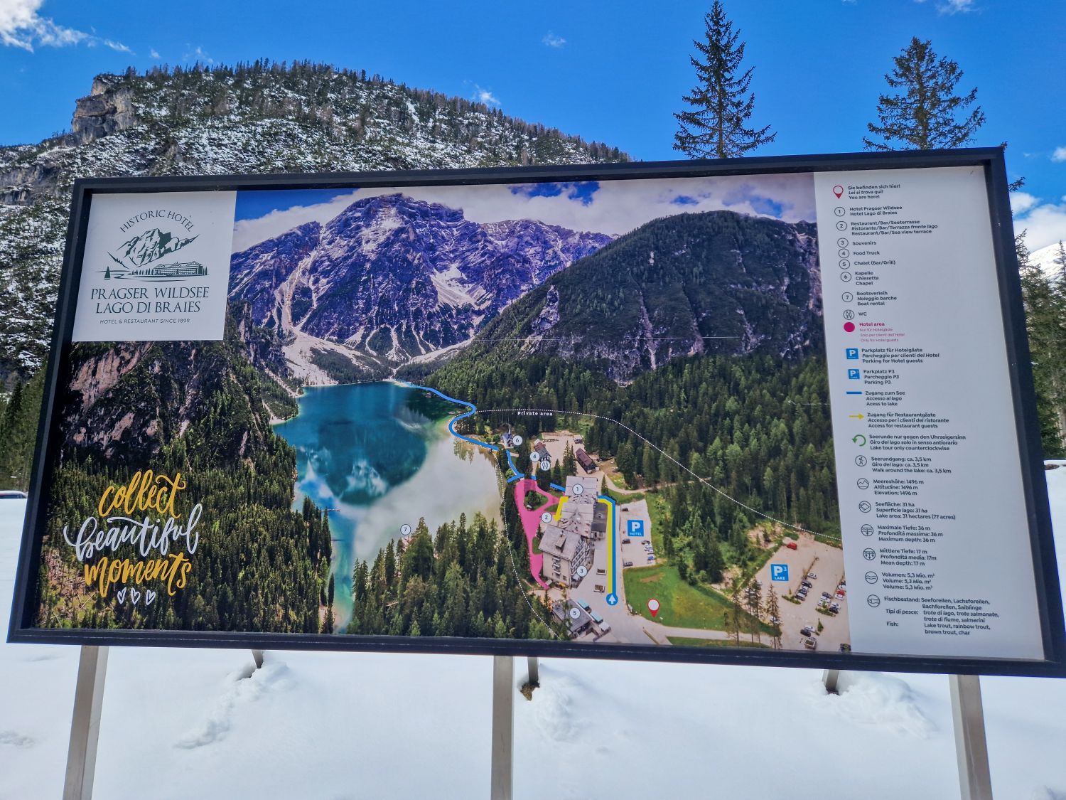 Lago di Braies: Discover The Pearl of the Dolomites 6 Large informational sign in a snowy landscape showing a map of Lago di Braies (Pragser Wildsee) with a photo of the lake, walking paths, buildings, legends, and the phrase “collect beautiful moments.” Pine trees and mountains are in the background.