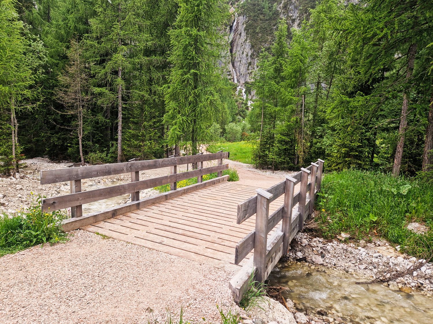 A wooden footbridge crosses a small stream in a forested area, surrounded by lush green trees and rocky terrain. A dirt path leads onto the bridge, with mountains visible in the background.