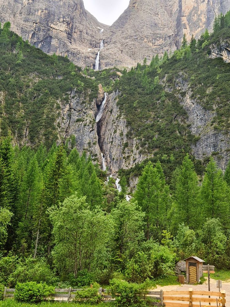 A tall, narrow waterfall cascades down a steep, rocky mountain surrounded by dense green pine trees and a wooden shed in the foreground.