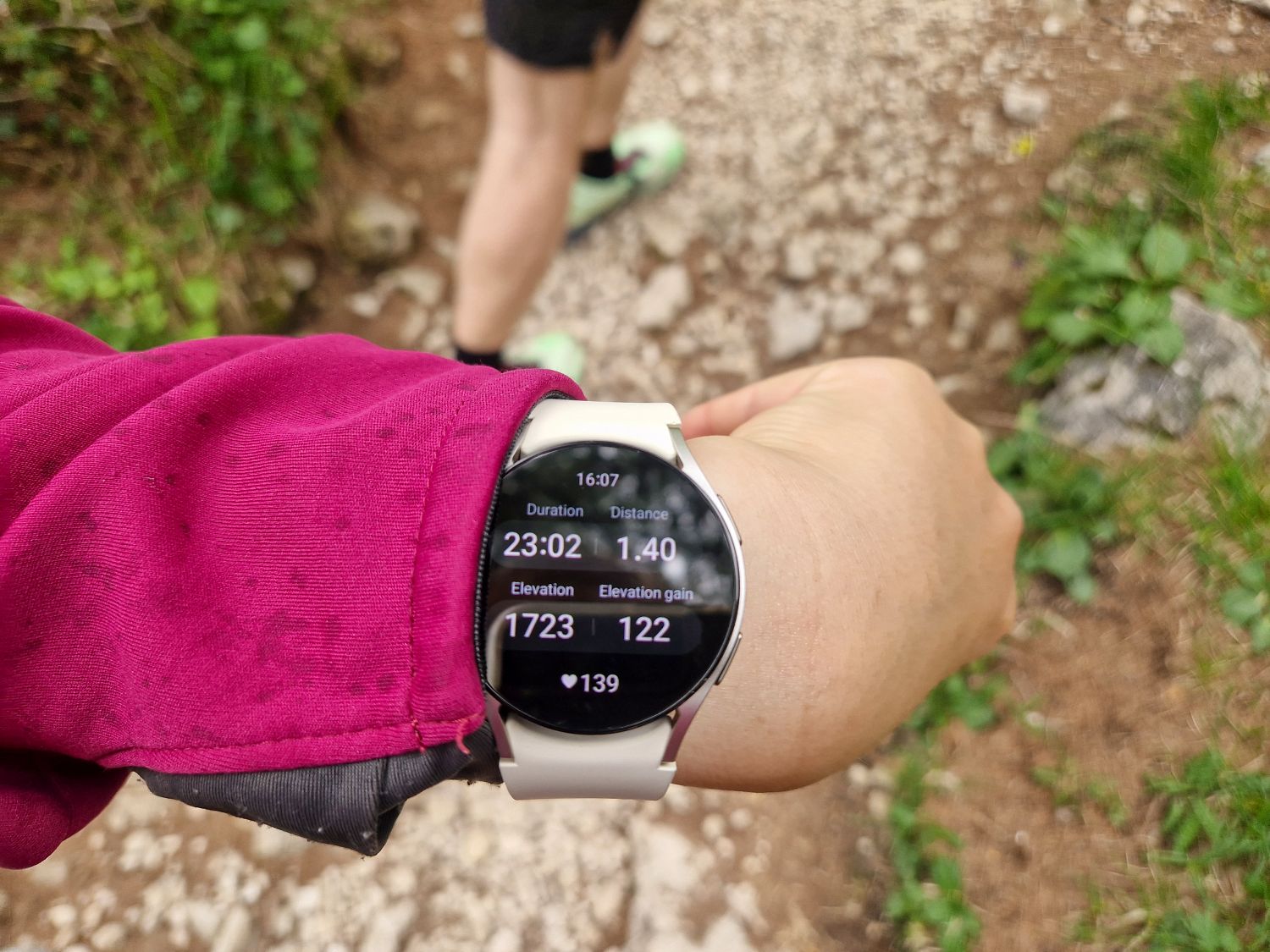 A person wearing a maroon jacket checks their smartwatch, displaying hiking stats: 23:02 duration, 1.40 km distance, 1723 m elevation, 122 m elevation gain, and 139 bpm heart rate. Another hiker walks ahead on a rocky trail.