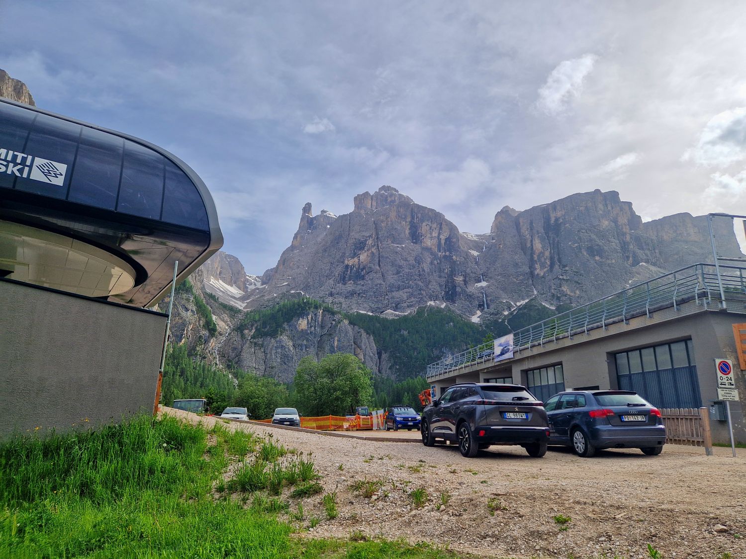 Cars parked near a modern ski lift station, with dramatic rocky mountains and green forests in the background under a partly cloudy sky.