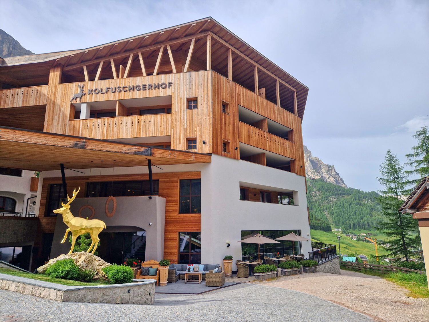 A modern wooden hotel with the name "Kolfuschgerhof" on the facade. There's a large golden deer statue on a rock near the entrance, with outdoor seating and mountains visible in the background.