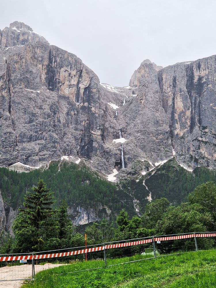 A tall, rocky mountain with patches of snow and a narrow waterfall cascading down the center, bordered by a green forest and a striped barrier in the foreground.