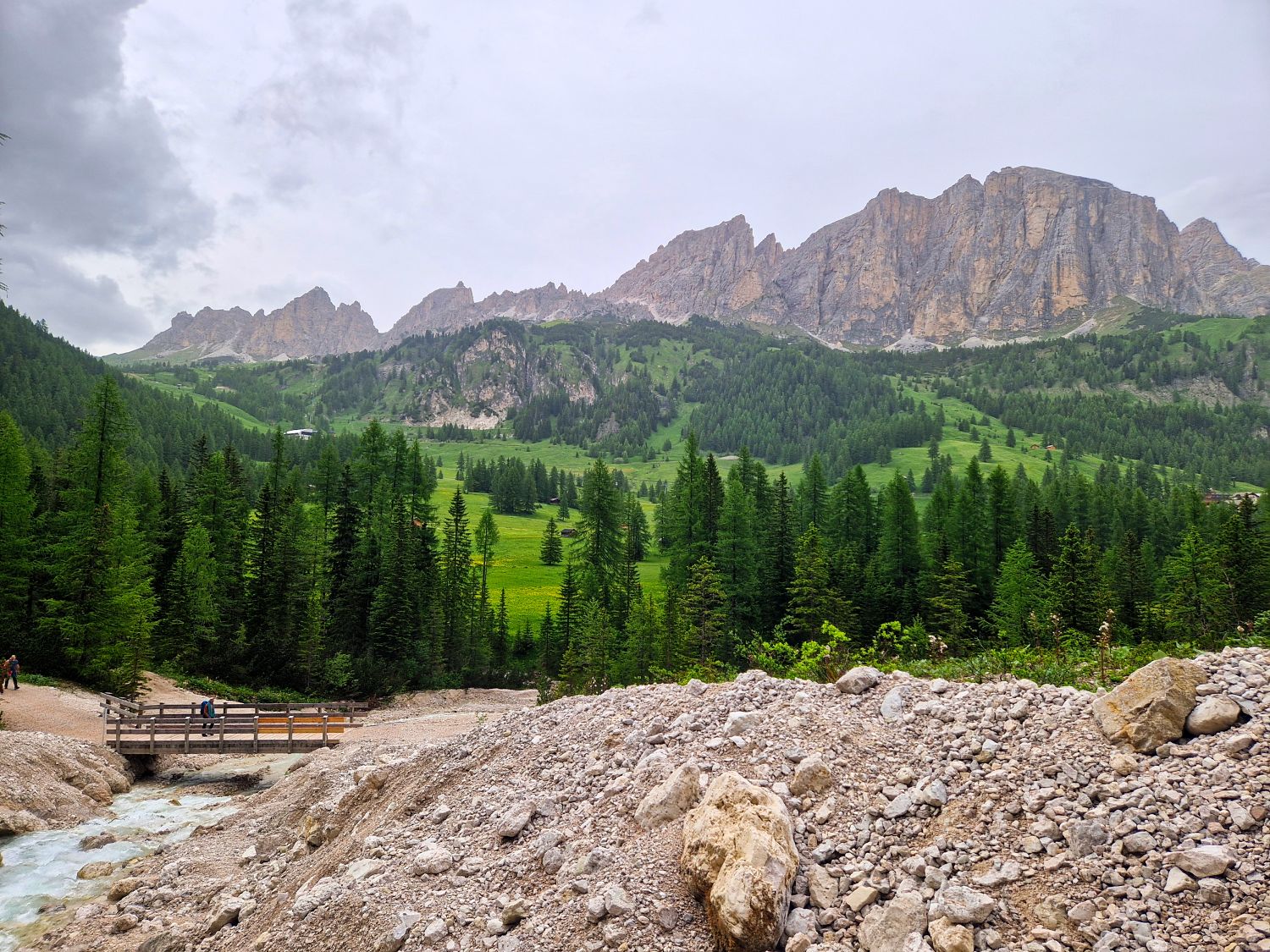 A rocky foreground leads to a small wooden bridge over a stream, surrounded by dense pine trees and green hills, with tall, rugged mountains in the background under a cloudy sky.
