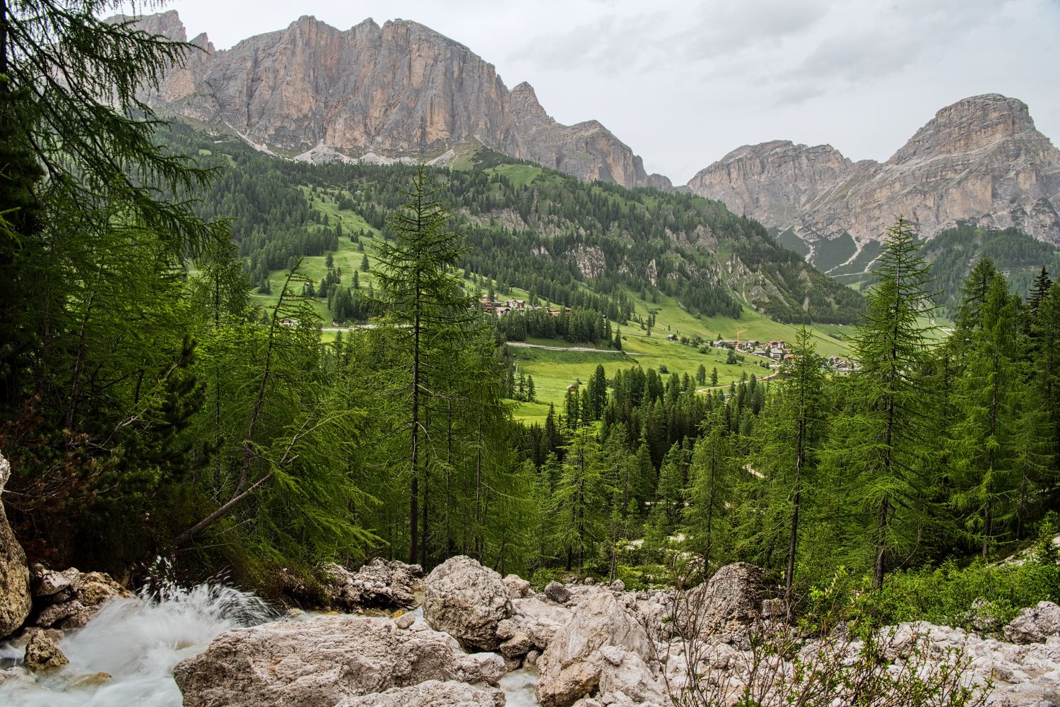 A lush, green valley with pine trees, rocky outcrops, and a small stream in the foreground, nestled beneath rugged, towering mountains under an overcast sky. Small houses are scattered in the valley.