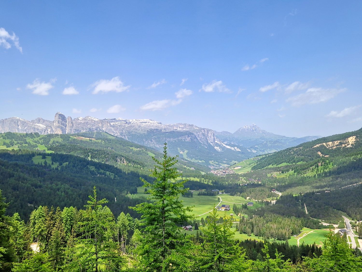 A scenic view of a lush green valley surrounded by forested hills and snow-capped mountains under a bright blue sky with scattered clouds. Small houses and fields are visible among the greenery.
