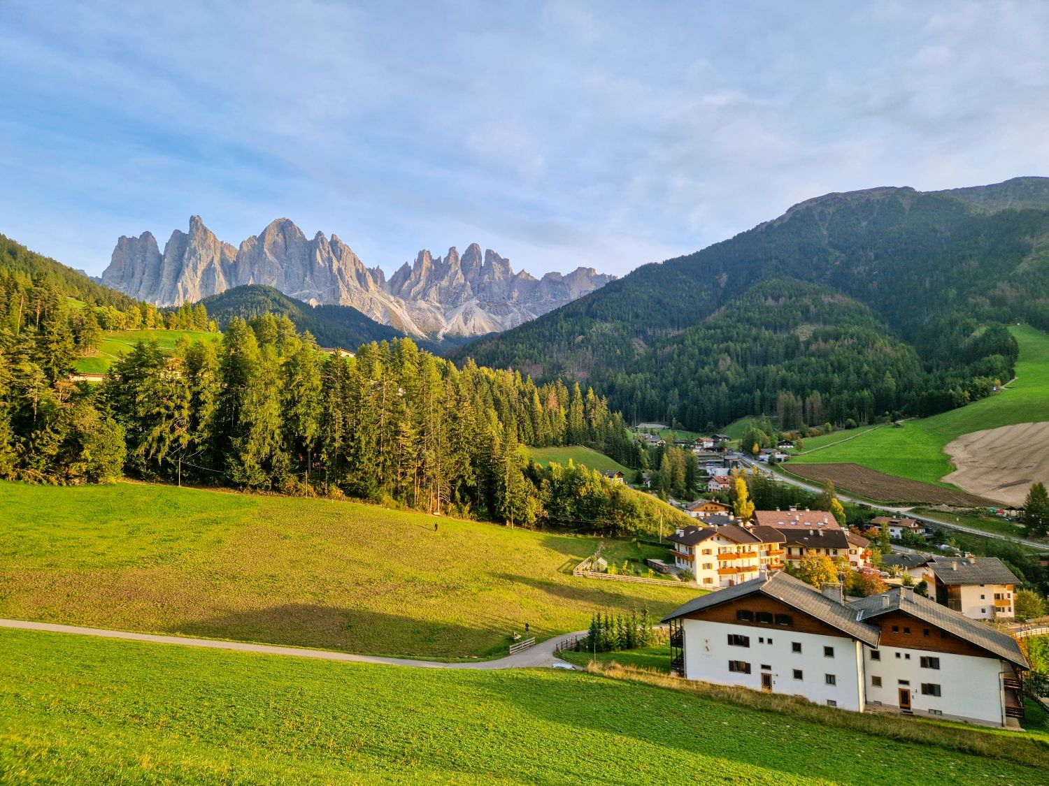 A picturesque village nestled in a green valley with forested hills, white houses, and dramatic, jagged mountain peaks in the background under a clear sky.
