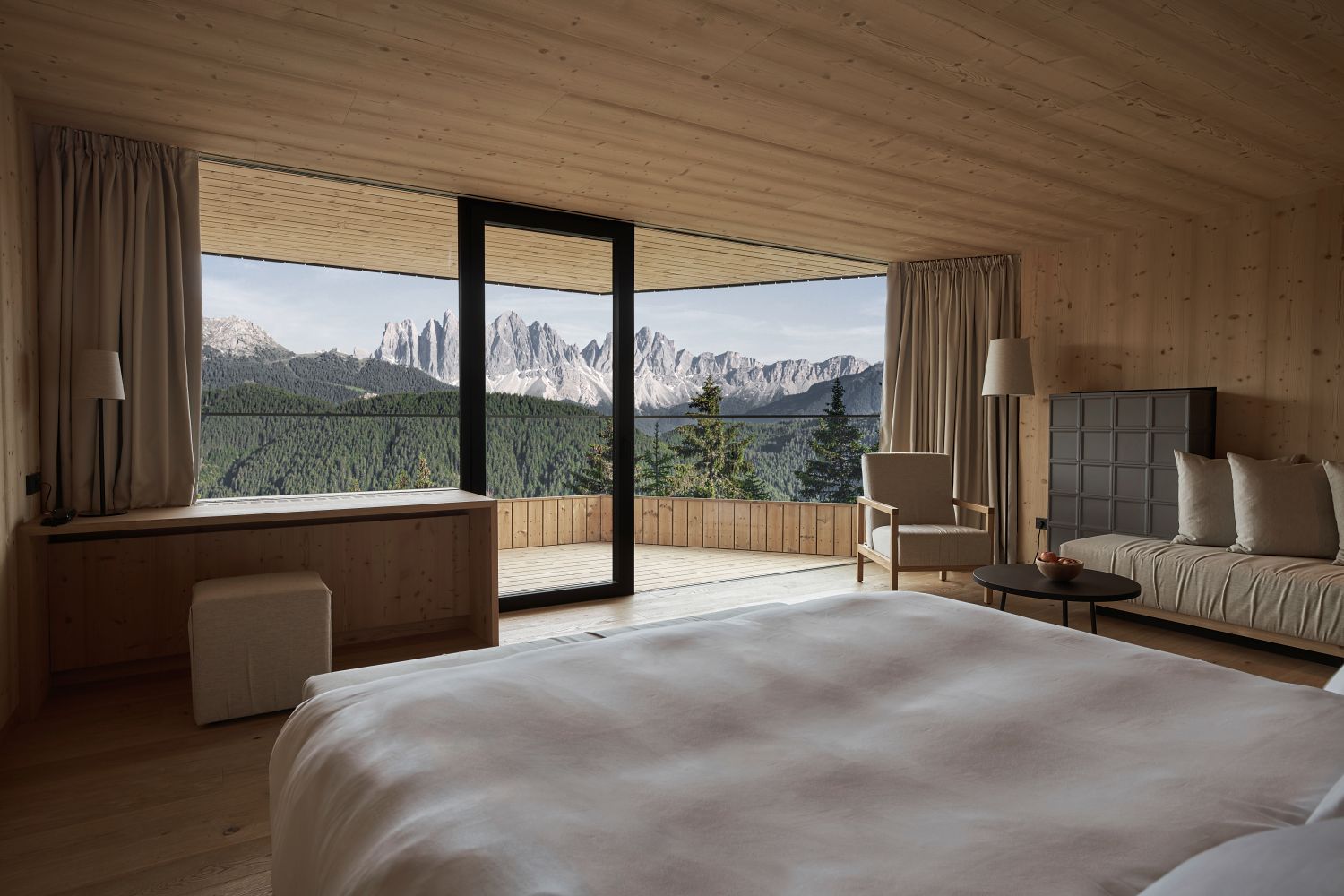 A modern, wood-paneled bedroom with a large bed, sofa, and floor-to-ceiling windows opening onto a balcony, offering a scenic view of green forests and distant mountains under a clear sky.