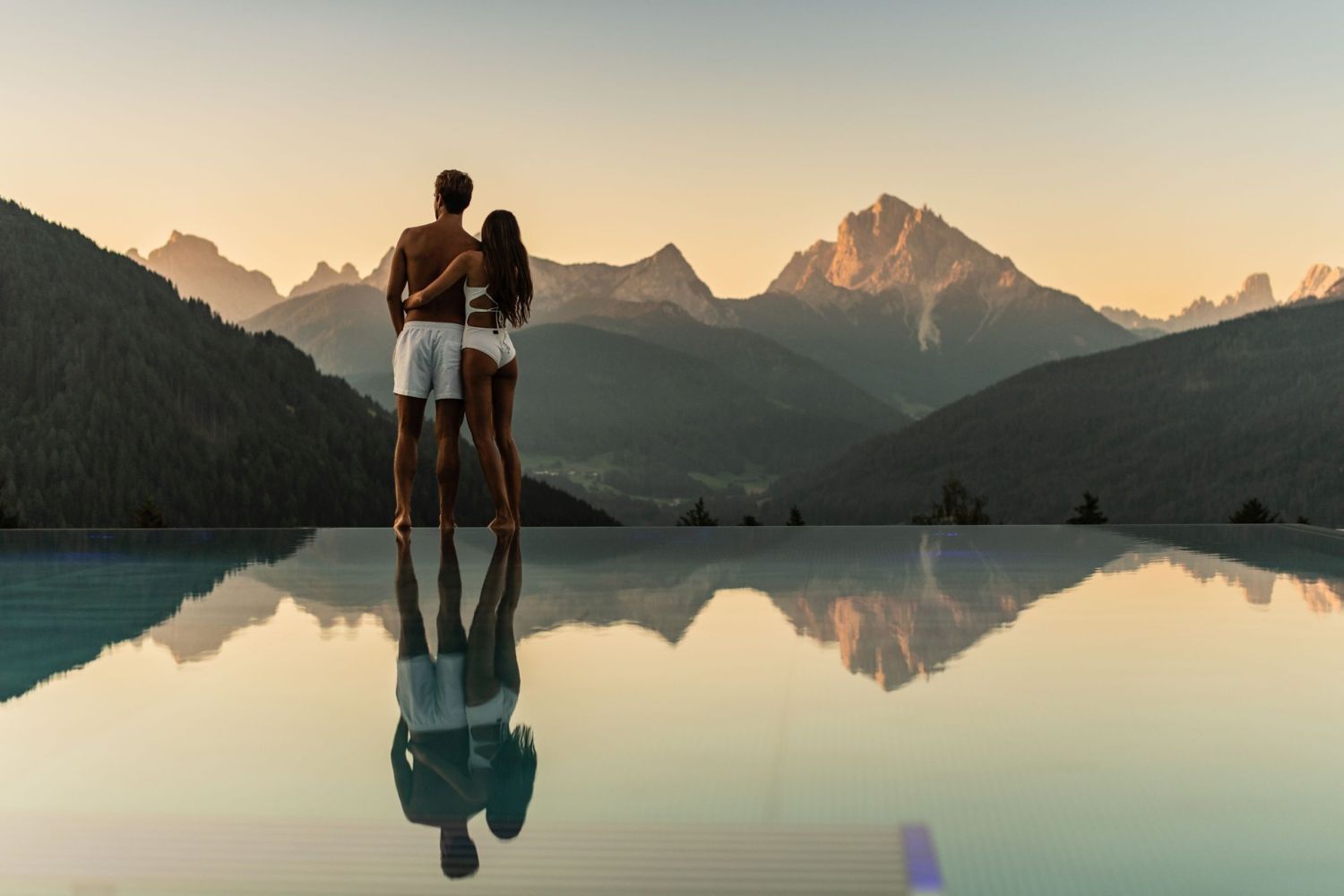 A couple in swimwear stands at the edge of an infinity pool, embracing as they look at distant mountains during sunset. The mountains and couple are reflected in the calm pool water.