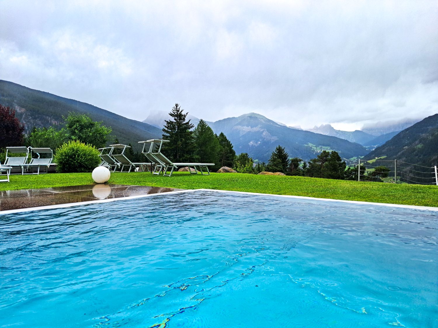 11 Hotels With The Best Views Of The Dolomites: Worth Every Penny 11 Outdoor pool at Hotel Scherlin in Ortisei with turquoise water rippling in the foreground. Lounge chairs sit on a rain damp lawn overlooking forested mountains and a misty alpine valley beneath dramatic gray clouds.