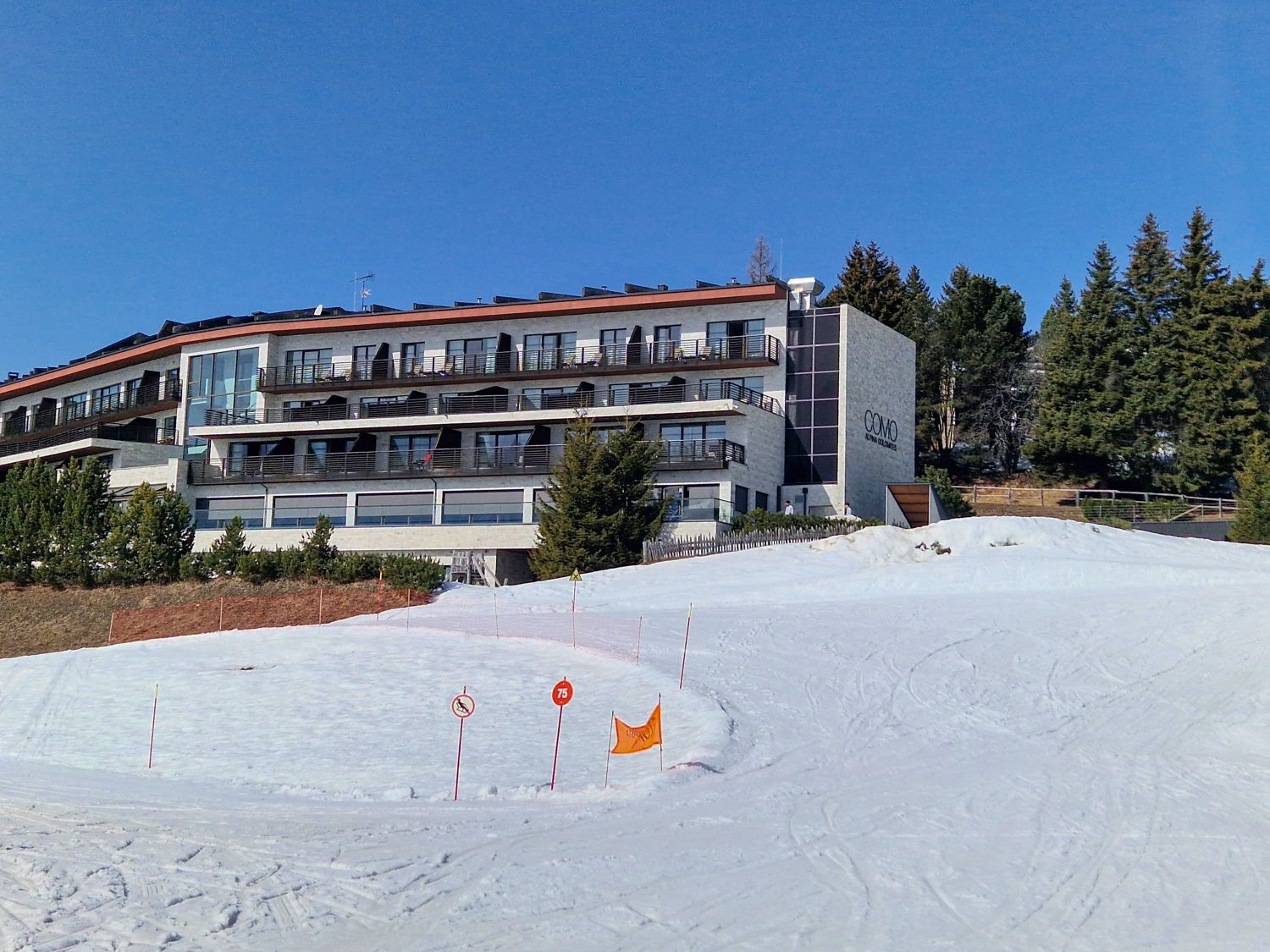 11 Hotels With The Best Views Of The Dolomites: Worth Every Penny 13 Modern multi story hotel building beside a snow covered ski slope under a clear blue sky. Pine trees line the hillside behind the hotel and orange trail markers stand in the snow, highlighting the property's direct access to the ski area.