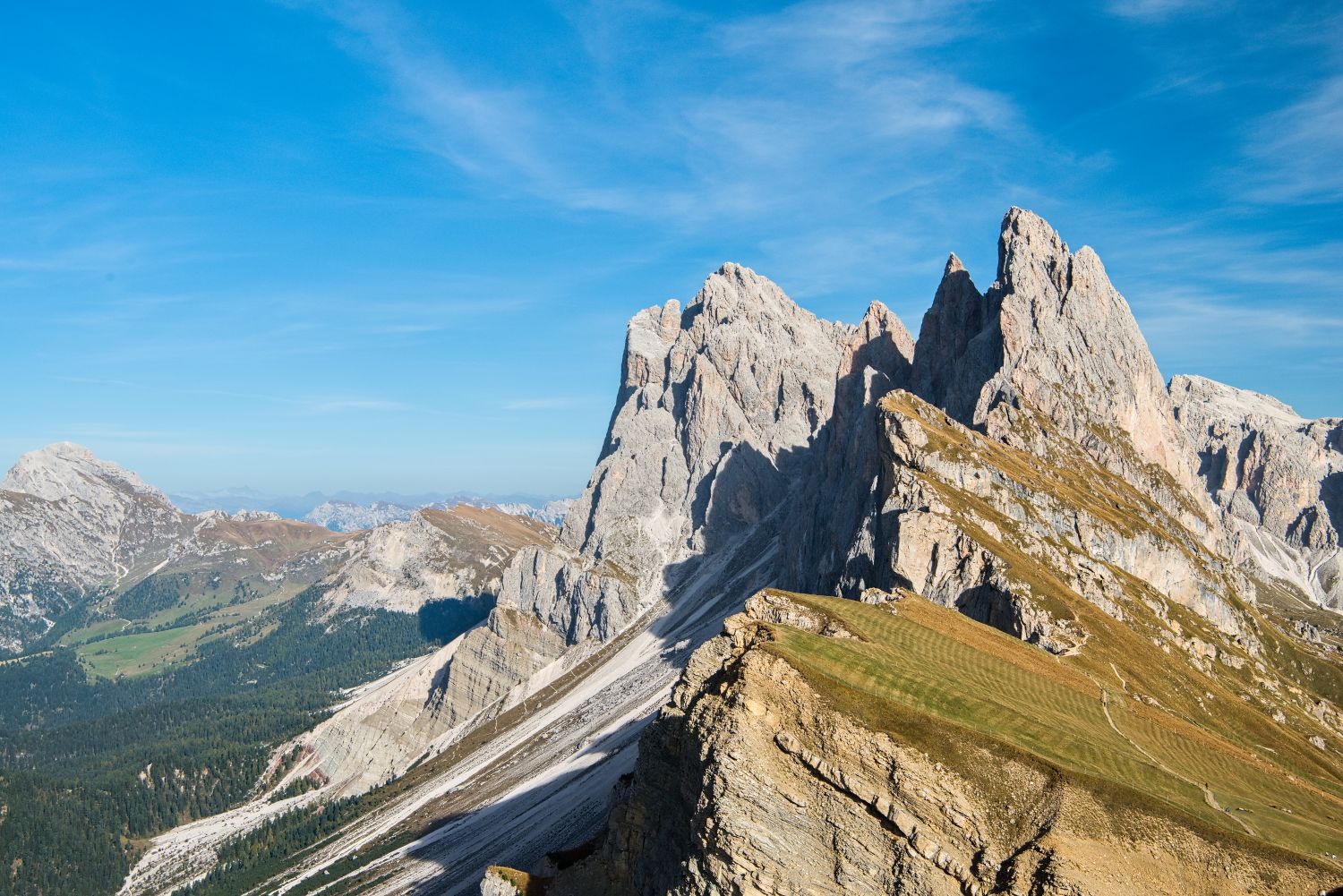 Mountain landscape with jagged rocky peaks, grassy slopes, and patches of snow under a clear blue sky. Rolling hills and distant mountains are visible in the background.