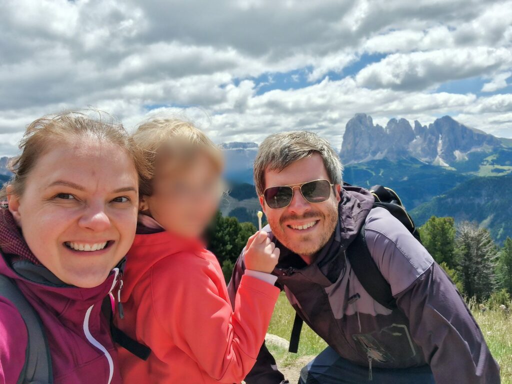 About 2 A smiling woman, a man in sunglasses, and a young child in outdoor jackets pose for a selfie on a grassy hillside with mountains and a partly cloudy sky in the background. The child's face is blurred.