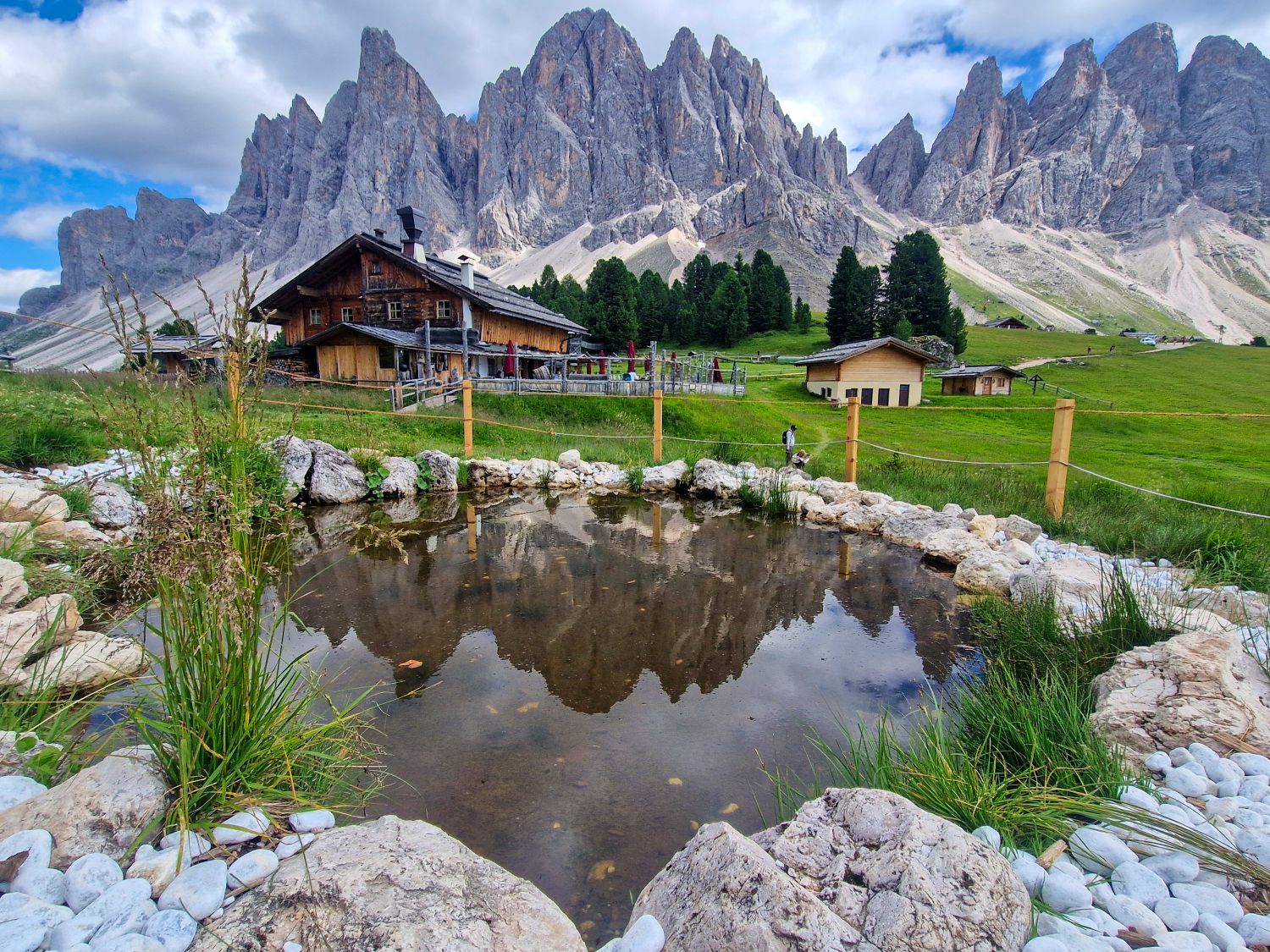 A rustic wooden house and smaller buildings sit on green grass near a rocky pond, with dramatic jagged mountains rising in the background under a partly cloudy sky.