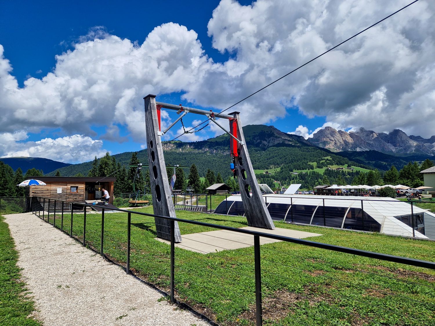 A zipline launch platform stands on a grassy hillside with a safety fence, a small wooden building nearby, and mountains under a partly cloudy sky in the background.