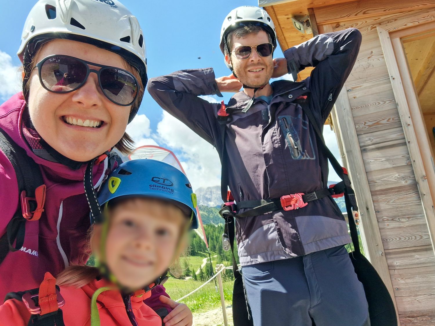 About 5 A smiling family of three wearing helmets and outdoor jackets stand together outside on a sunny day, ready for an adventure. A wooden building and green landscape are visible in the background.