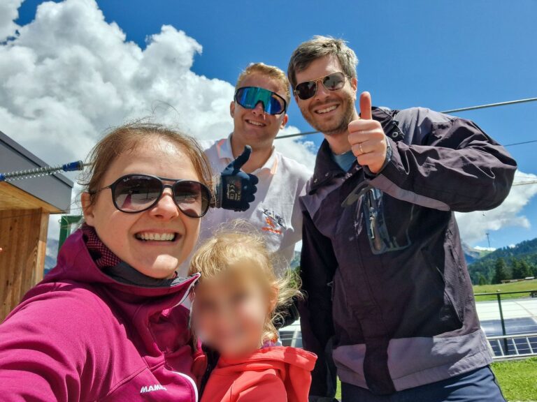 Four people pose outdoors on a sunny day, three adults with sunglasses smiling at the camera and giving thumbs up. A child in front is wearing a red jacket, with their face blurred. Green hills and blue sky are in the background.