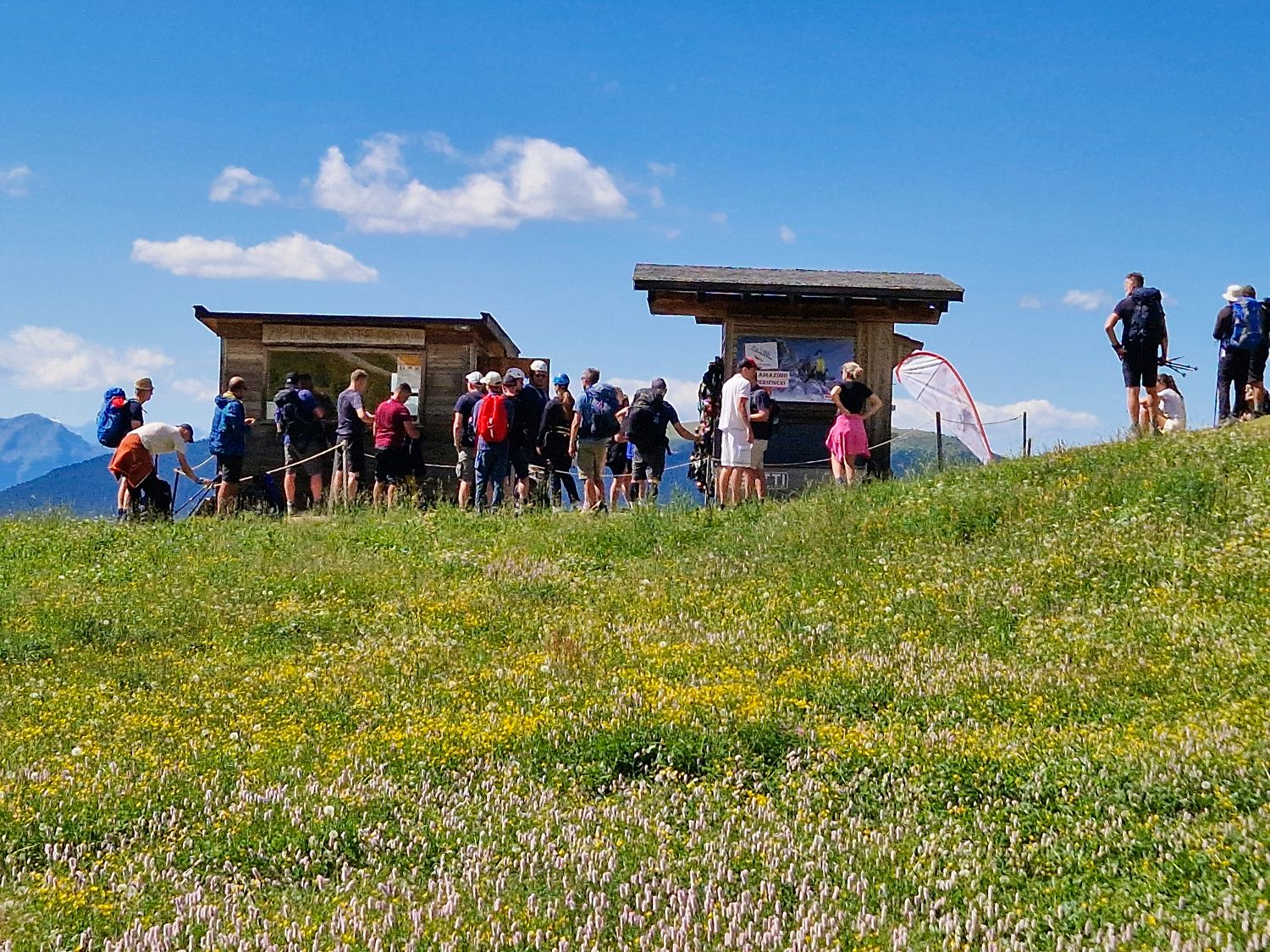 Visitors gather at a mountain viewpoint on a grassy alpine hillside dotted with wildflowers under a bright blue sky. People stand near information boards and a launch area at Zipline Monte Pana, capturing the busy atmosphere of this popular outdoor attraction in the Dolomites.