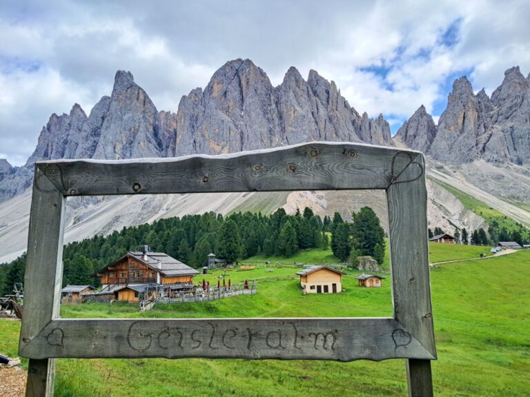 Wooden frame with "Geisleralm" carved on it, overlooking alpine huts, green meadows, pine trees, and jagged mountain peaks under a cloudy sky in a picturesque mountain valley.