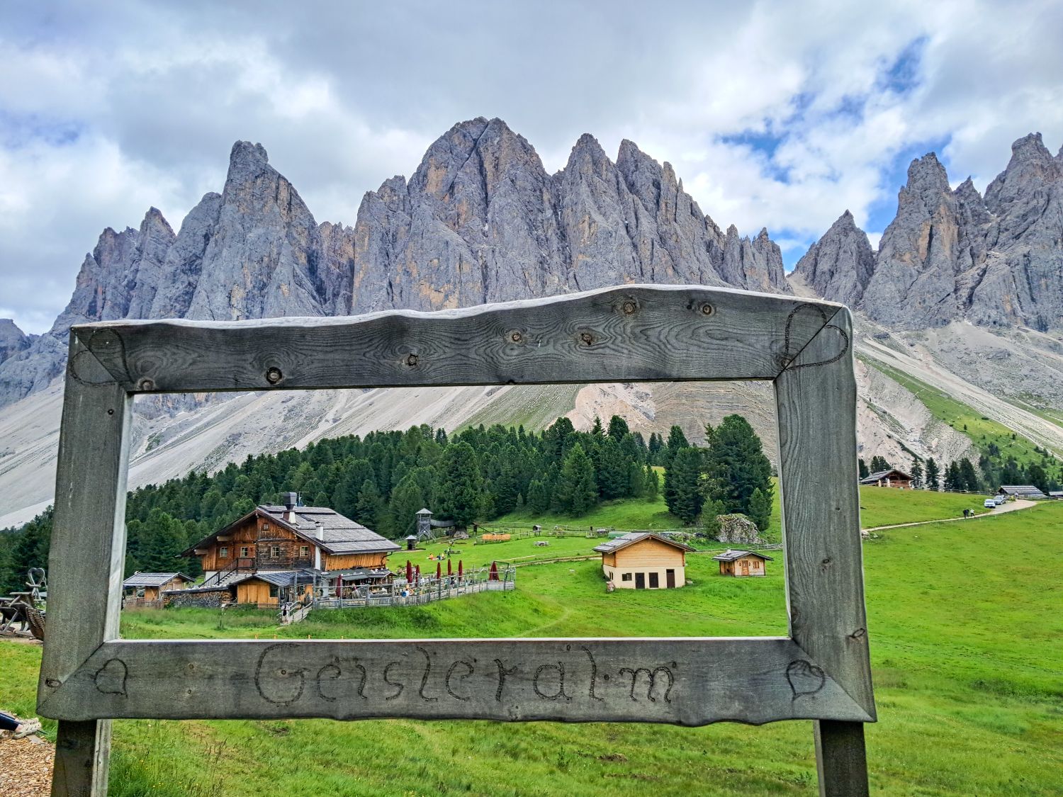 Wooden frame with "Geisleralm" carved on it, overlooking alpine huts, green meadows, pine trees, and jagged mountain peaks under a cloudy sky in a picturesque mountain valley.