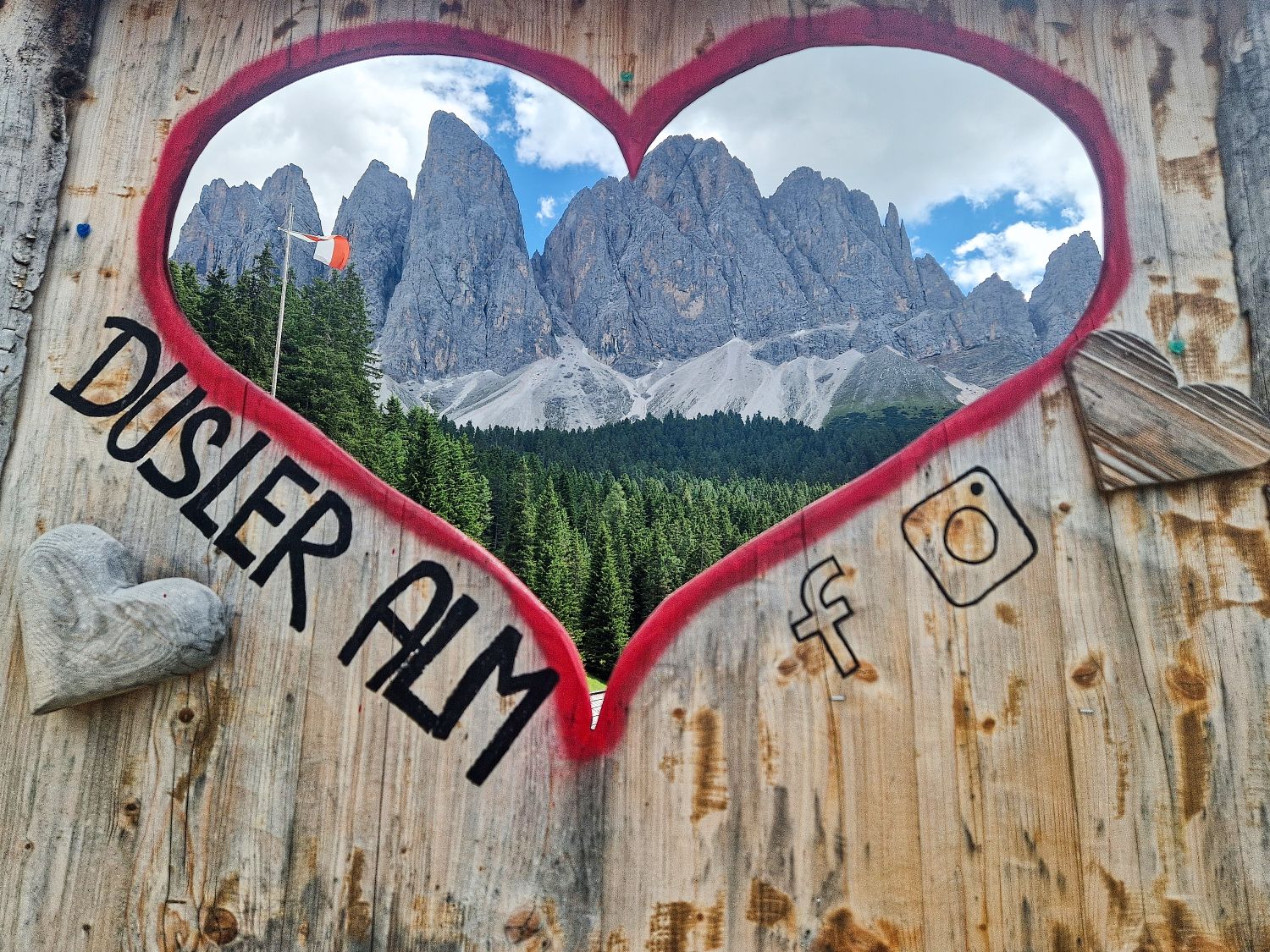 Wooden frame with a red heart outline and "Dusler Alm" text, looking out onto tall, jagged mountain peaks, a green forest, and a partly cloudy sky. Social media icons and wooden heart decorations are on the frame.