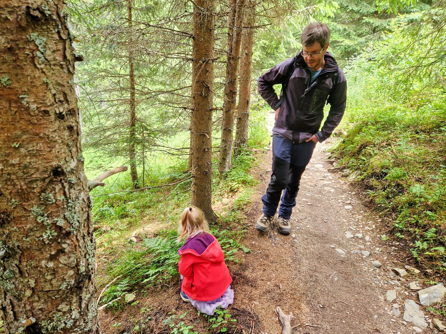 Home 13 A man walks on a forest trail while a young child in a red jacket sits on the ground nearby, surrounded by green trees and foliage.
