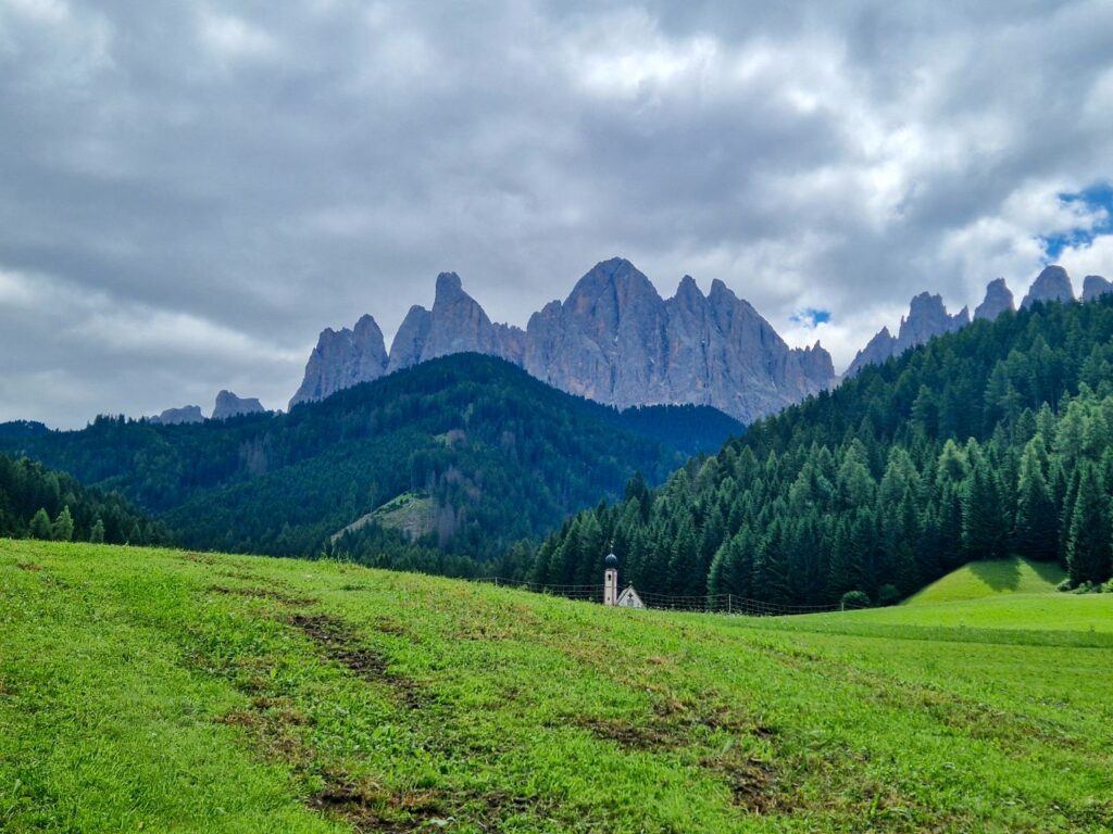 Green meadow with tire tracks in the foreground, a small white chapel among dense pine trees, and dramatic rugged mountains under a cloudy sky in the background.