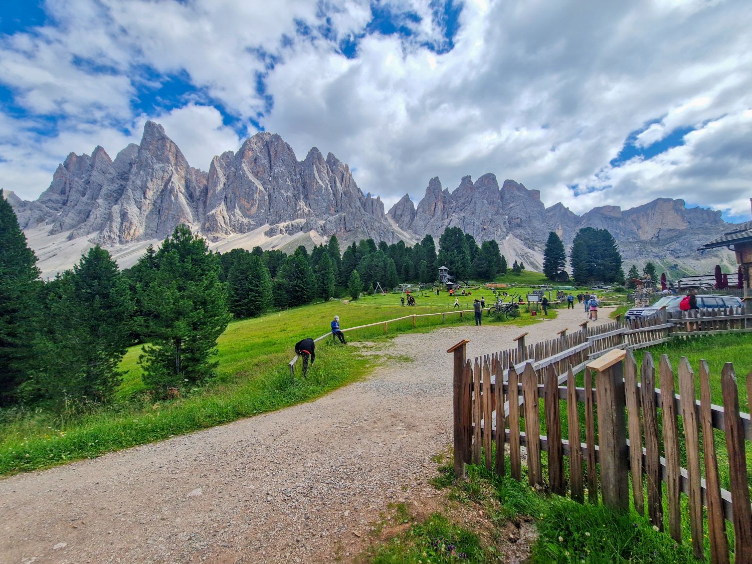 A scenic mountain landscape with jagged peaks, lush green grass, pine trees, a gravel path, and several people walking or relaxing outdoors under a partly cloudy sky. A wooden fence lines the right side of the image.