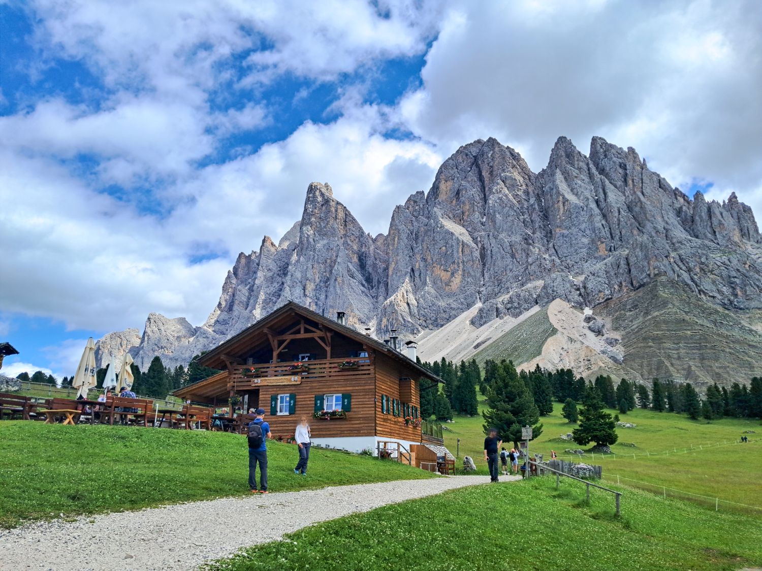 A wooden cabin sits on a grassy hill with people walking nearby. Tall, jagged mountains rise dramatically in the background under a partly cloudy blue sky. Pine trees dot the landscape.