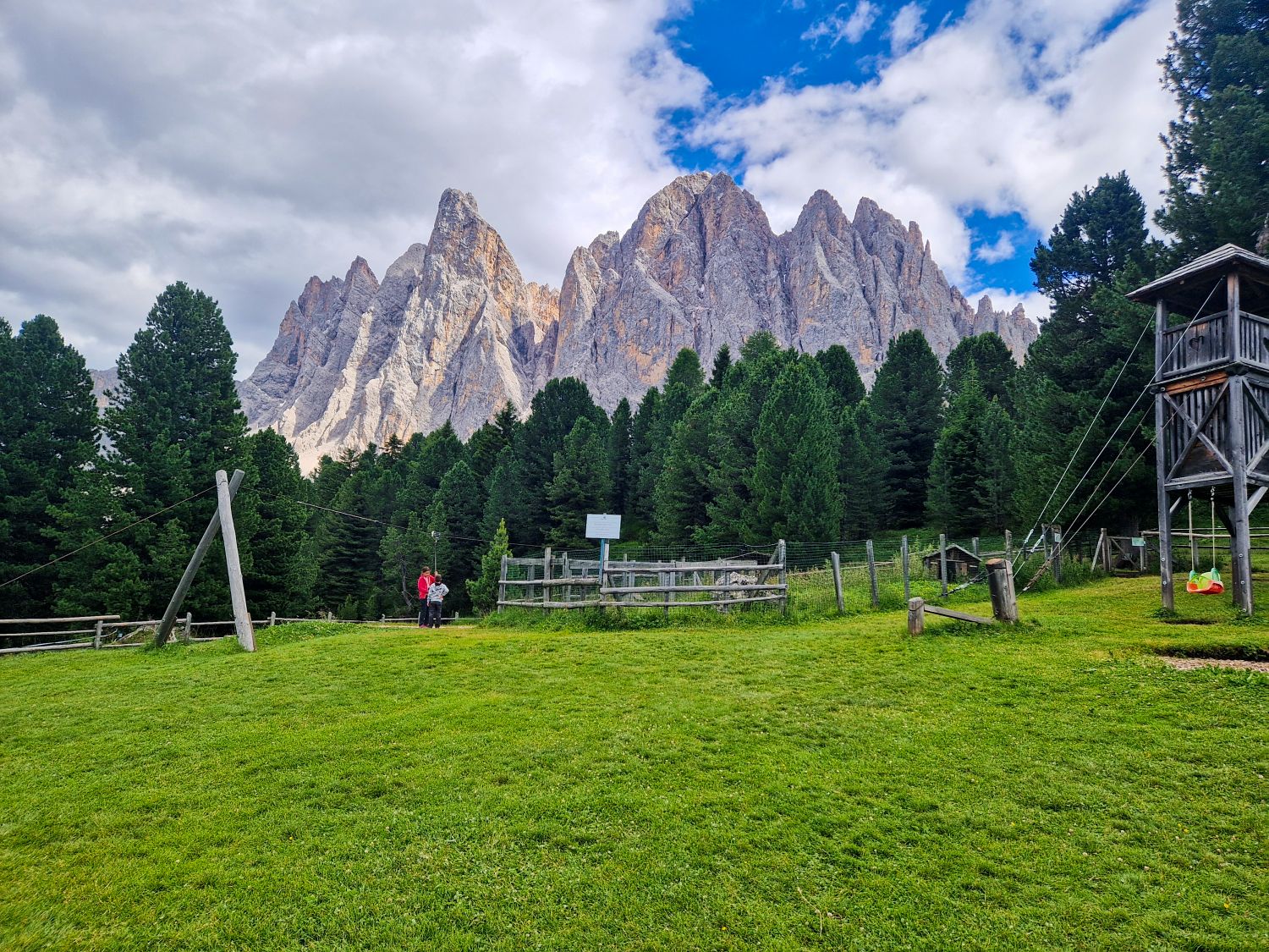 A lush green field with wooden playground equipment and fences, bordered by dense pine trees. Jagged mountain peaks rise dramatically in the background under a partly cloudy sky. Two people stand near the fence.
