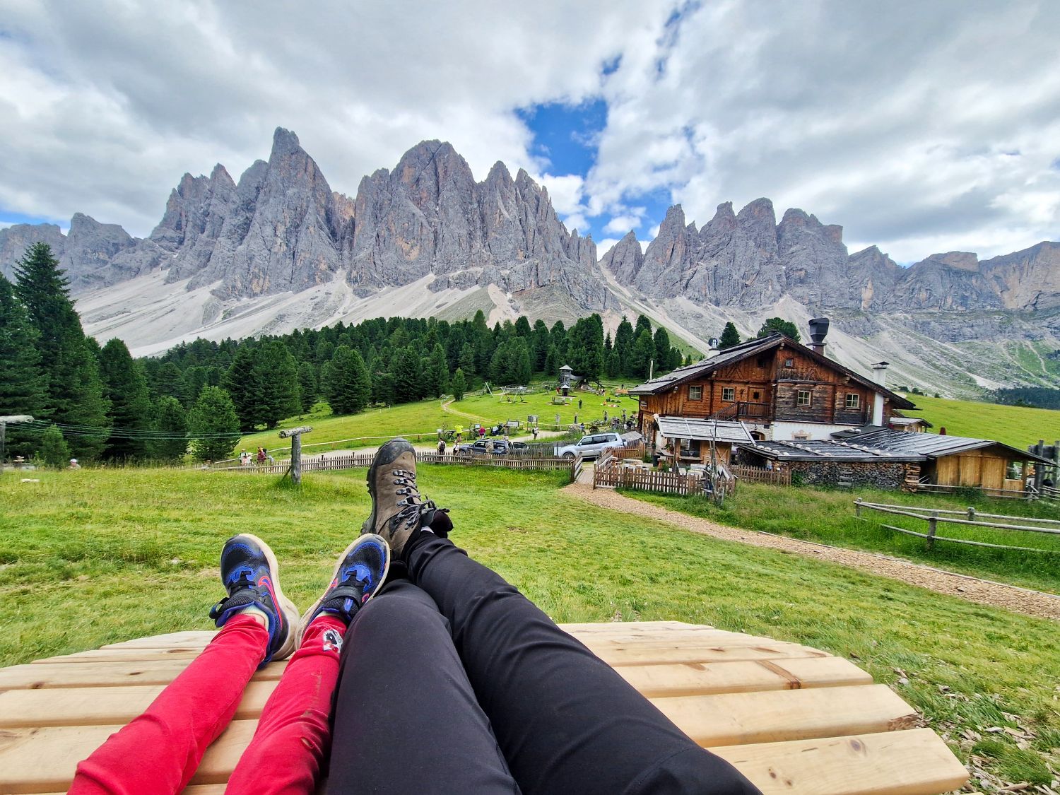 Home 12 Two people lying on a wooden bench in a grassy field with a rustic cabin, pine trees, and dramatic mountain peaks in the background under a partly cloudy sky.