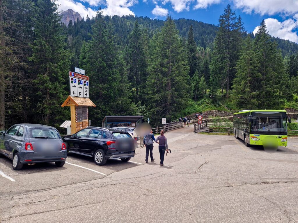 A small parking lot in a forested area with parked cars, a green bus, two people walking, and direction signs; tall pine trees and mountains are in the background under a partly cloudy sky.