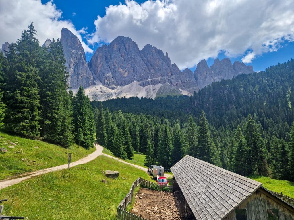 A wooden cabin and fenced area sit among green trees and grassy hills, with a dirt path winding through a forest. Jagged mountain peaks rise dramatically in the background under a partly cloudy sky.