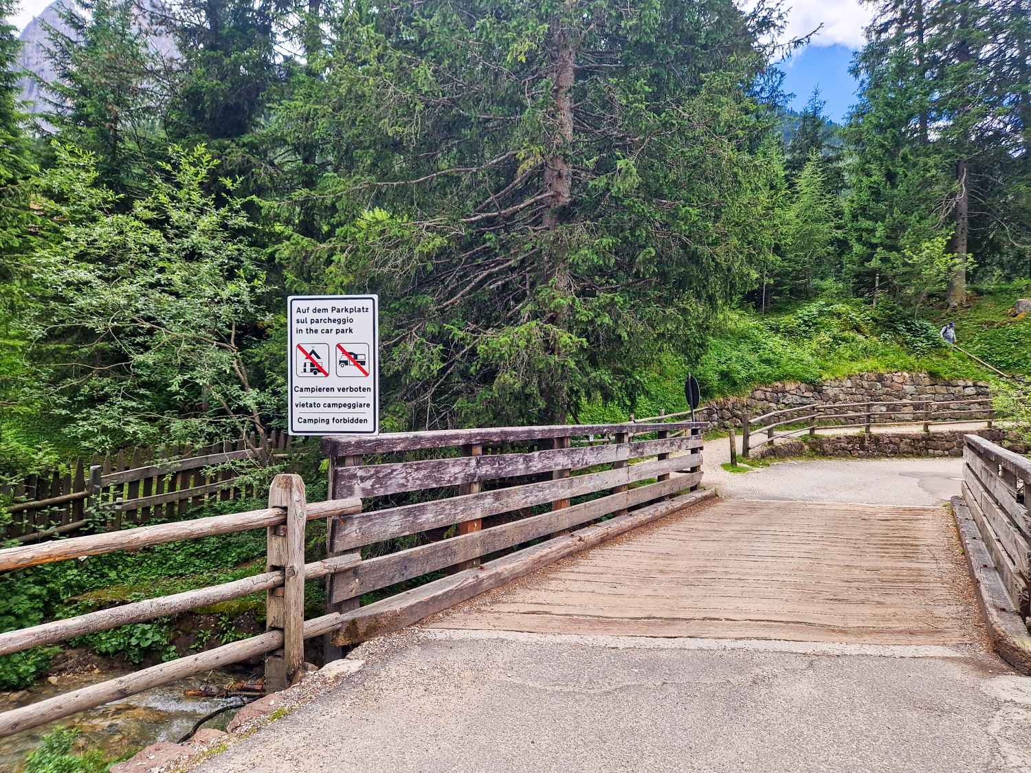 A wooden bridge surrounded by green trees with a sign prohibiting camping and open fires. The path continues into a forested area under a partly cloudy sky.
