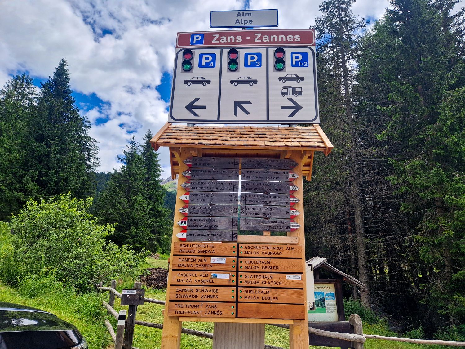 A wooden signpost in a forested area shows parking directions for cars and motorhomes at Zans–Zannes with arrows and icons, as well as hiking trail names and destinations below on wooden plaques.