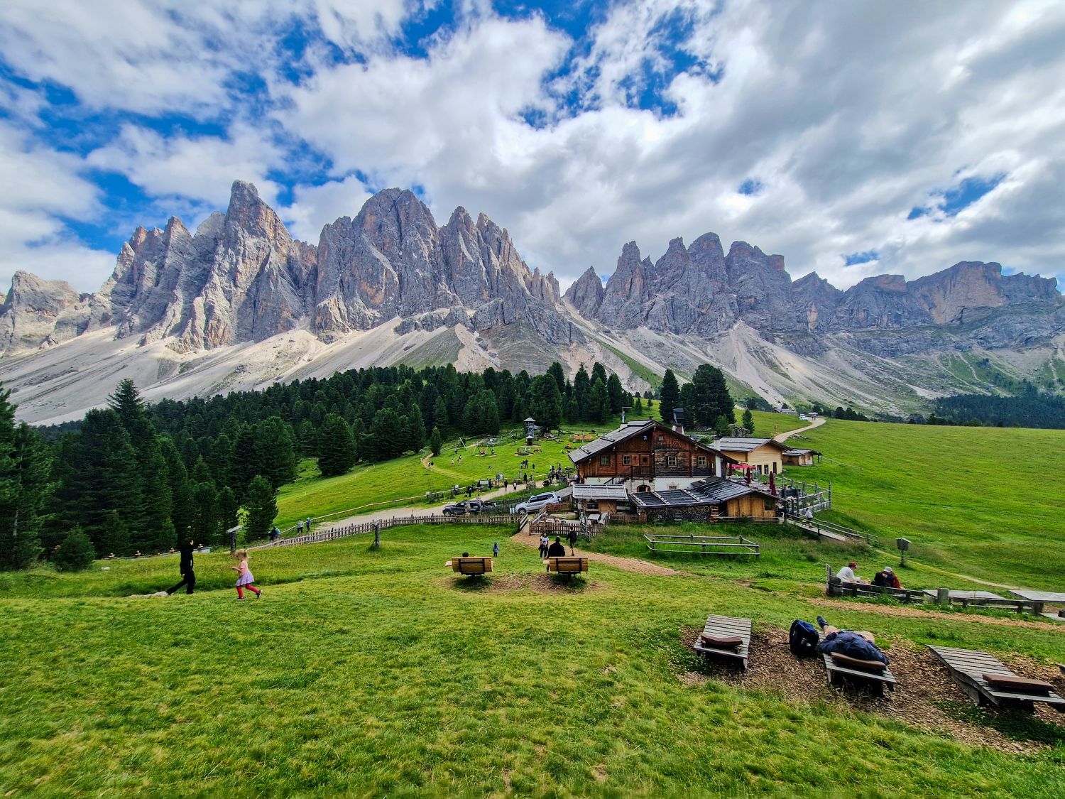 Chalet surrounded by green grass and pine trees with dramatic jagged mountains in the background under a partly cloudy sky; people are walking and relaxing outdoors.