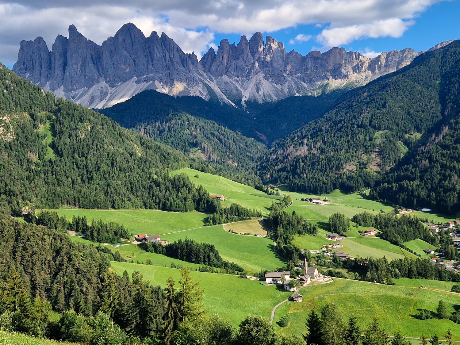 A lush green valley with scattered houses and a church is surrounded by dense forests and dramatic, jagged mountains beneath a partly cloudy sky.