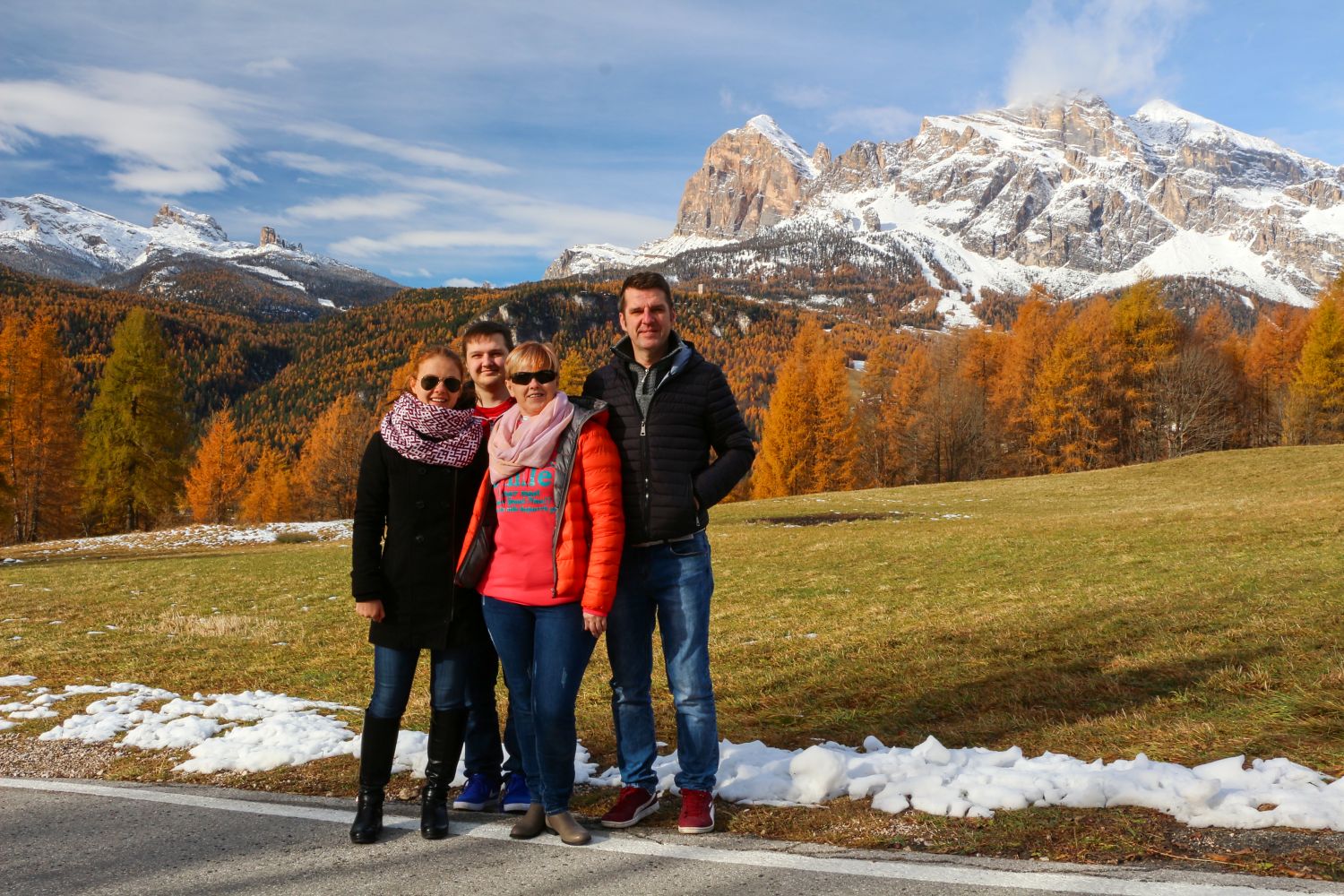 Four people stand together outdoors in front of snow-dusted mountains and autumn-colored trees, wearing jackets and scarves, enjoying a sunny day with patches of snow on the ground.